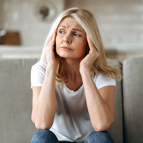 Middle-aged woman sits on a couch, hands pressed to her temples, looking worried or stressed in thought.
