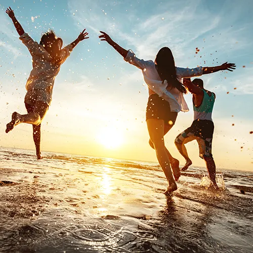 Three people jump with arms raised on a wet beach at sunset, water droplets spraying around them.
