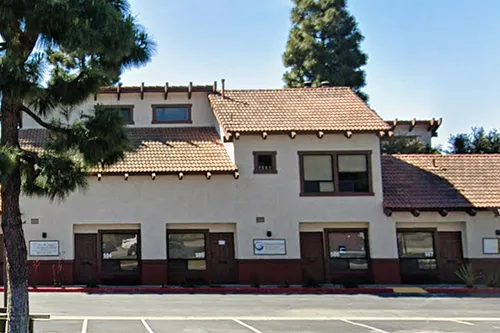 Two-story beige stucco building with a red-tile roof, several windows and exterior signs, next to a parking lot with a tree in the foreground.