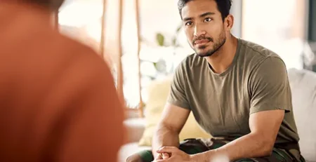 Man in an olive T-shirt and camo pants sits on a couch, facing a blurred person, listening intently in a living room.