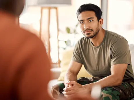Man in an olive T-shirt and camo pants sits on a couch, facing a blurred person, listening intently in a living room.