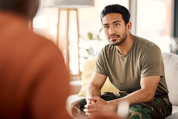 Man in an olive T-shirt and camo pants sits on a couch, facing a blurred person, listening intently in a living room.