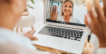 Person on a video call waves hello from a laptop screen during a video chat. Close-up shows the user's hand on the table with the screen displaying a smiling woman.