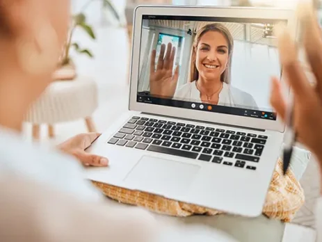 Person on a video call waves hello from a laptop screen during a video chat. Close-up shows the user's hand on the table with the screen displaying a smiling woman.