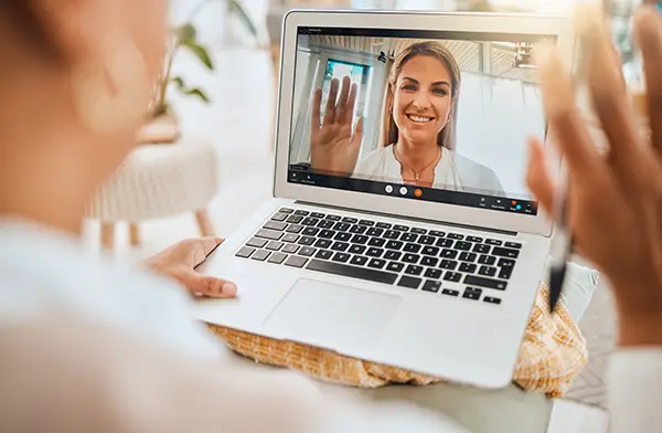 Person on a video call waves hello from a laptop screen during a video chat. Close-up shows the user's hand on the table with the screen displaying a smiling woman.