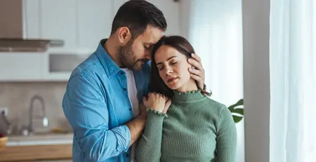 A man comforts a sad woman in a bright kitchen, holding her head and shoulder as they stand by a window at home.