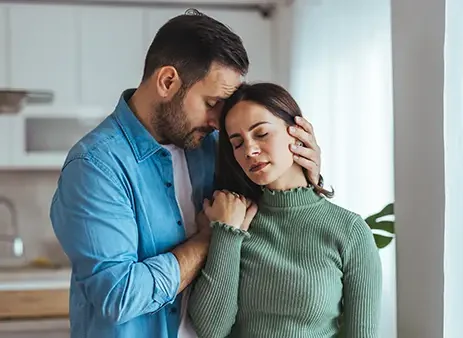 A man comforts a sad woman in a bright kitchen, holding her head and shoulder as they stand by a window at home.