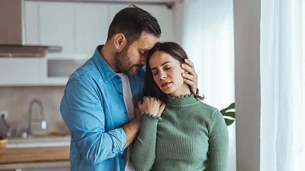 A man comforts a sad woman in a bright kitchen, holding her head and shoulder as they stand by a window at home.